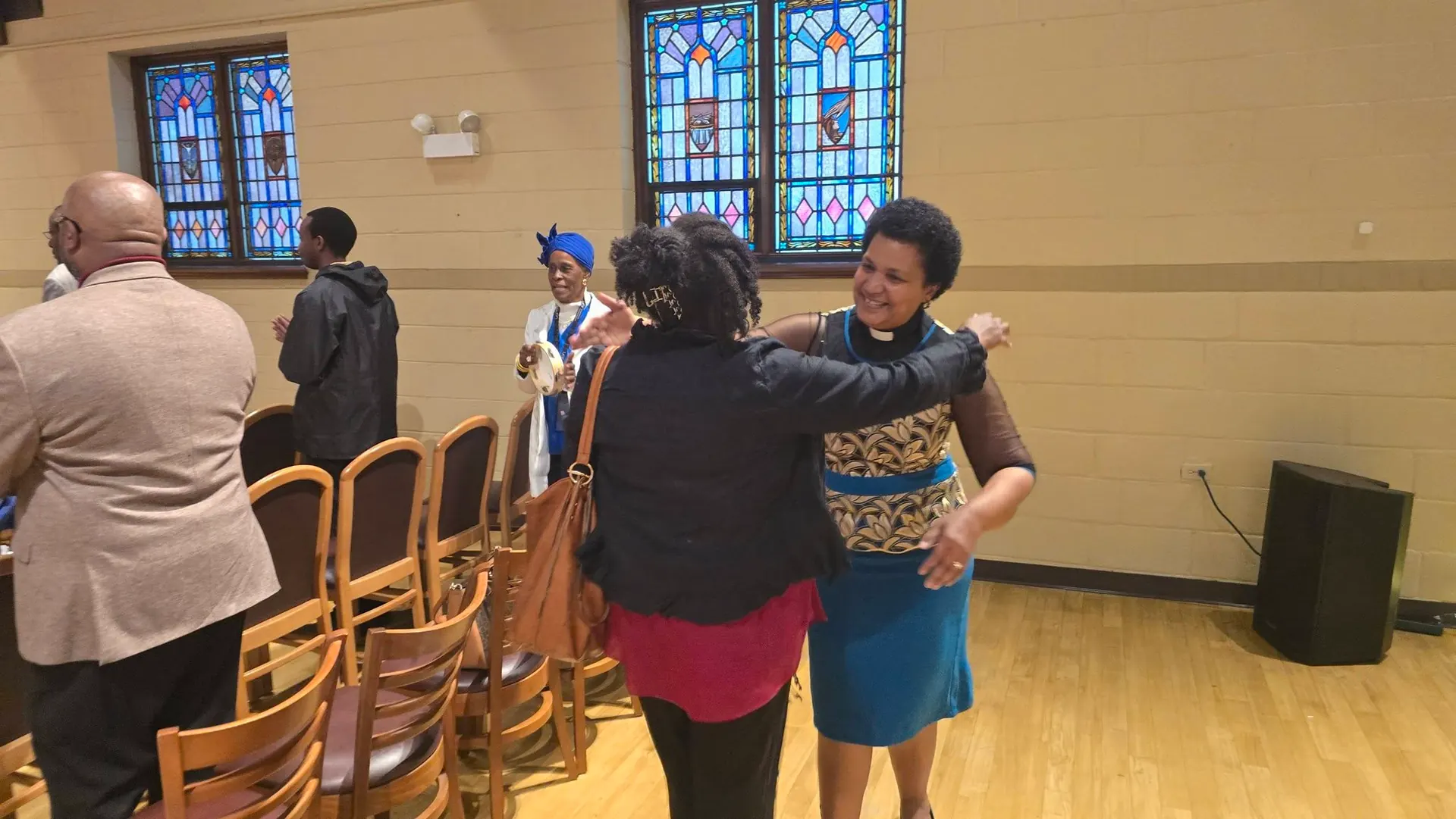A pastor and a member of Tumaini Swahili Chapel embrace in the sanctuary after worship, with other members talking behind them